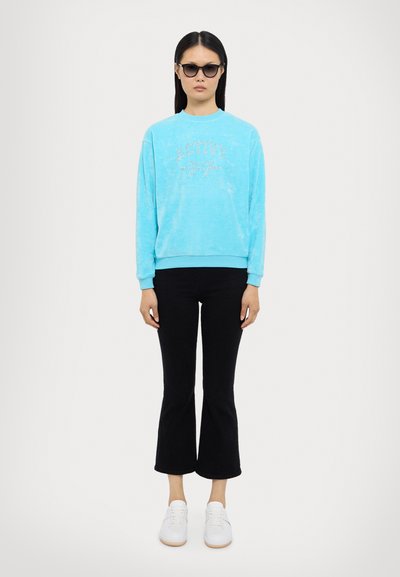 Lyseblå velourgenser med "ACTIVE" i rhinestones, kombinert med svarte flagrende bukser og hvite sneakers. Stylet med svarte solbriller.