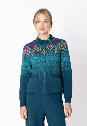 Junge Frau trägt eine geknöpfte, türkisfarbene Strickjacke mit mehrfarbigen Blumen- und geometrischen Mustern über passenden türkisfarbenen Hosen.