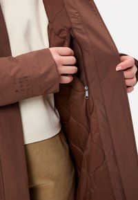 Veste matelassée marron avec une poche zippée, présentant une texture lisse et un design ajusté. Les manches affichent un texte imprimé près du poignet.