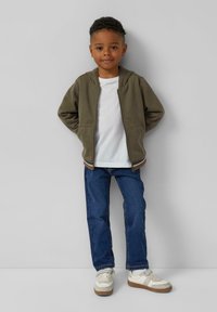 Olivově zelená mikina na zip, bílý tričko, modré džíny a bílé tenisky s hnědými detaily. Jednoduchý design a volný střih.