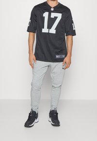 Person iført en sort NFL-trøje med nummer 17, lysegrå Nike sweatpants og sorte og hvide Nike sneakers, stående foran en ensfarvet baggrund.