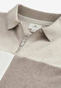 Polo pour homme à fermeture éclair en beige, avec un panneau blanc texturé et un tissu beige lisse. Comprend un col et un détail de fermeture éclair argentée.