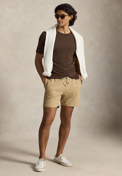Άνδρας με γυαλιά ηλίου, καφέ κοντομάνικο πουκάμισο, μπεζ σορτς, λευκά αθλητικά παπούτσια και λευκή ζακέτα περασμένη στους ώμους, στέκεται με τα χέρια στις τσέπες.