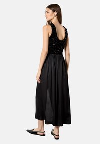 Schwarzes Maxikleid mit einem Pailletten-Oberteil, ärmellosem Design und einem fließenden Rock mit leichtem Glanz. Der Rücken hat einen runden Ausschnitt.