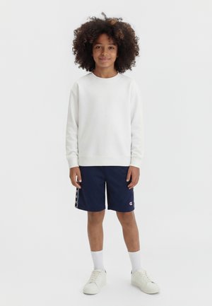 Dziecko z kręconymi włosami, ubrane w białą bluzę z długim rękawem, granatowe szorty, białe skarpetki i białe trampki, stojące na jednolitym tle.
