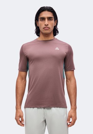 Ung mand med mørkt hår iført en mauve kortærmet træningsshirt og lysegrå shorts, stående foran en ensfarvet baggrund.