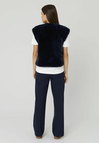 Gilet en fausse fourrure marine avec un col montant, des épaules larges et une texture lisse ; porté par-dessus un T-shirt blanc et associé à un pantalon sombre.