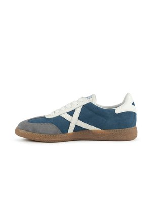 Zapatilla baja con ante azul, puntera gris, cordones blancos y diseño lateral en X, con suela de goma marrón texturizada.