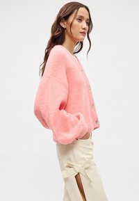 Ροζ, αφράτη ζακέτα με V-λαιμόκοψη, μακριά φουσκωτά μανίκια και κλείσιμο με τέσσερις κουμπιά. Φοριέται πάνω από ανοιχτόχρωμα παντελόνια με πλευρικές σχισμές και κορδόνια.