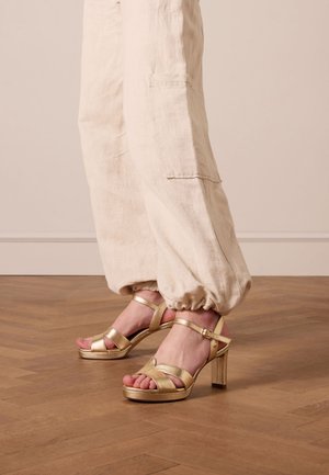 Sandali con tacco alto in metallo dorato, dotati di plateau e cinturino alla caviglia. Presentano cinturini incrociati e un tacco a blocco spesso. Sfondo in legno.