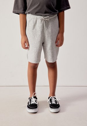 Enfant portant un short en polaire gris clair, des baskets noires avec des lacets blancs, et un t-shirt à manches courtes gris foncé, debout sur un fond uni.
