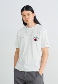 Weißes Baumwoll-T-Shirt mit dem "Champion"-Logo in Rot und Schwarz auf der linken Brust, Rundhals, kurze Ärmel, lockere Passform.