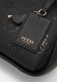 Sac à main noir texturé Guess avec logos floraux embossés, étiquette attachée avec "GUESS EST 1981" en lettres dorées, et petite clé décorative.