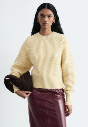 Femme aux longs cheveux noirs portant un pull côtelé crème, une jupe en cuir bordeaux, des boucles d'oreilles créoles dorées et tenant un sac marron foncé dans sa main gauche.