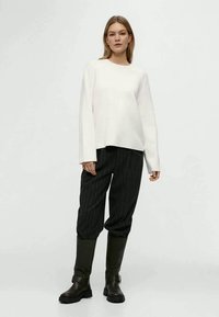 Donna che indossa un top lungo bianco largo, pantaloni neri a righe e stivali neri sopra il ginocchio, in piedi contro uno sfondo semplice.