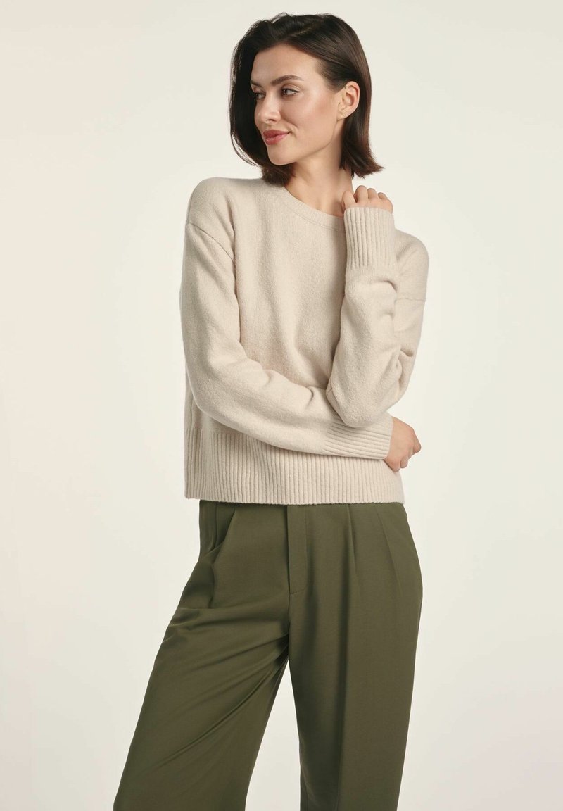 Beżowy sweter z dzianiny z prążkowanymi mankietami i dołem, noszony z luźnymi oliwkowozielonymi spodniami. Miękka tekstura, luźny krój, okrągły dekolt.