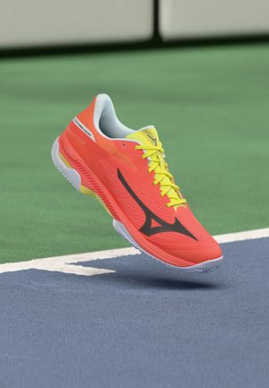 Chaussure de sport orange vif et jaune avec logo noir flottant au-dessus d'un court de tennis près de la ligne.
