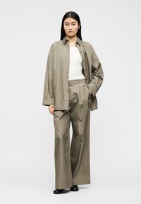 Γυναίκα με μακριά μαύρα μαλλιά, φορώντας χαλαρό πουκάμισο σε χρώμα ταουπέ με κουμπιά, φαρδιά παντελόνια στο ίδιο χρώμα, λευκή μπλούζα και μαύρα παπούτσια, στέκεται με το ένα της χέρι στην τσέπη.