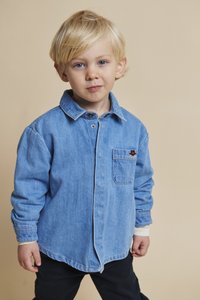 Lys blå denimskjorte med knapper, lange ermer, brystlomme og brodert detalj. Brukt med mørke bukser, mot en beige bakgrunn.