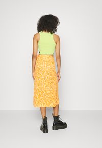 Top verde sem mangas e saia midi laranja com padrões ondulados. Botas de combate pretas com atacadores. Vista traseira, fundo simples.