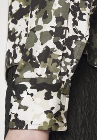 Blusa de manga longa em tecido leve, apresentando um padrão de camuflagem em tons de verde, preto e creme, com detalhes em botões dourados nos punhos.
