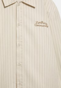 Chemise crème boutonnée avec des rayures verticales marron. Elle présente un col classique et un logo brodé "Northern Community" en marron.