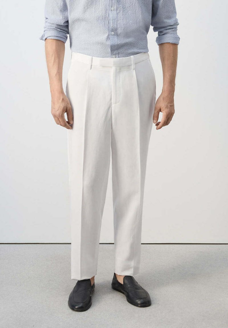 Homme portant un pantalon blanc plissé, une chemise bleu clair à manches longues retroussées et des chaussures noires sans lacets, debout sur un sol gris.