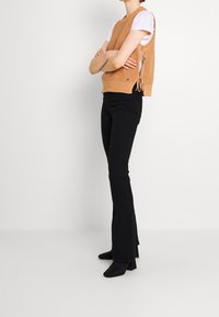 Gebreide mouwloze top in camel, met zijstrikken, gedragen over een witte T-shirt, in combinatie met zwarte flared broeken en enkellaarsjes met hak.