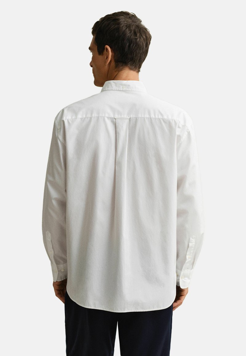Man die van achteren is gezien, draagt een wit overhemd met lange mouwen en een plooi aan de achterkant, samen met een donkere broek, tegen een effen achtergrond.