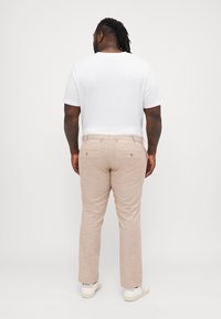 Helle beige Leinenhose mit strukturiertem Finish, ausgestattet mit zwei Gesäßtaschen mit goldenen Akzenten, kombiniert mit einem schlichten weißen T-Shirt.