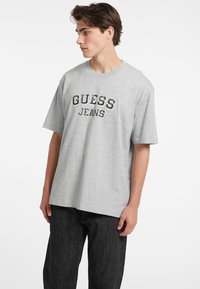 Pilkos spalvos medvilnės marškinėliai laisvu kirpimu, su užrašu "GUESS" ir "JEANS", didelėmis juodomis raidėmis išspausdintais ant krūtinės. Paprastas dizainas, trumpomis rankovėmis.