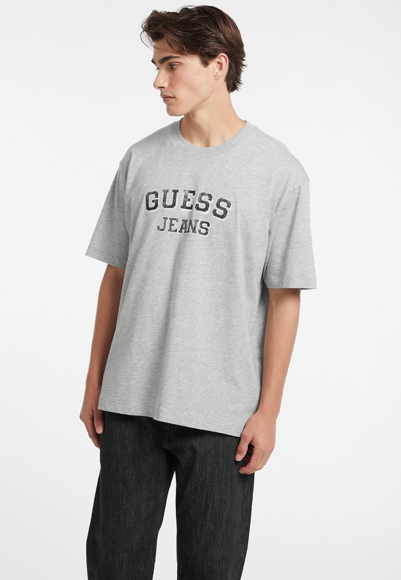 Pilkos spalvos medvilnės marškinėliai laisvu kirpimu, su užrašu "GUESS" ir "JEANS", didelėmis juodomis raidėmis išspausdintais ant krūtinės. Paprastas dizainas, trumpomis rankovėmis.