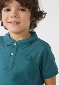 Jongen met krullend haar draagt een teal polo shirt met een klein JOTT-logo op de borst, terwijl hij vriendelijk glimlacht tegen een effen achtergrond.