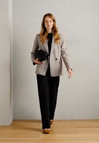Blazer à carreaux noir et blanc, pull noir et pantalon noir. Tenant un casque noir, avec un sol en bois clair et un fond gris.