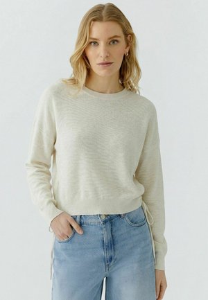 Vrouw met blond haar die een lichtbeige gebreide trui en lichtblauwe high-waisted spijkerbroek draagt, staand met één hand in de zak.