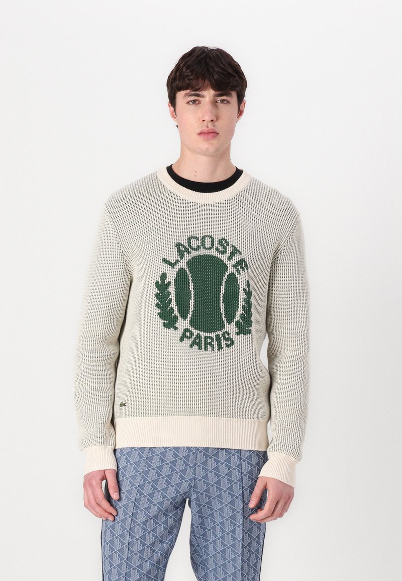 Pulover tricotat bej deschis, cu un logo brodat verde „LACOSTE PARIS” ce include un design cu o minge de tenis. Manșete și tiv cu arici.