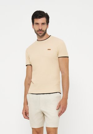 Uomo in piedi che indossa una camicia beige a maniche corte con bordo nero e pantaloncini beige abbinati su sfondo bianco.