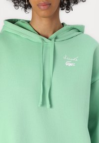 Žalias džemperis su minkšta medžiaga, su priekinėmis kengūros kišenėmis. Su baltu Lacoste logotipu ir reguliuojamomis raištelėmis ant gobtuvo.