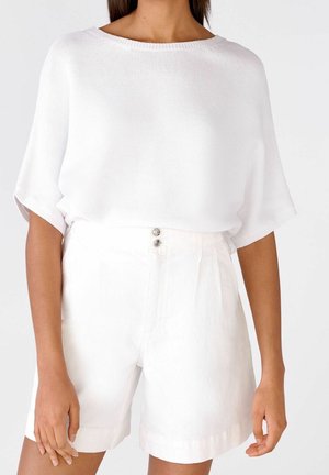 Kobieta nosząca luźny biały krótki top z krótkim rękawem oraz białe szorty z wysokim stanem z dwoma guzikami na pasie.