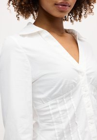 Γυναίκα που φοράει εφαρμοστό λευκό πουκάμισο με κουμπιά, βαθύ V-λαιμόκοψη και πιέτες στη μέση, μπούκλες μαλλιών ορατές στις άκρες.