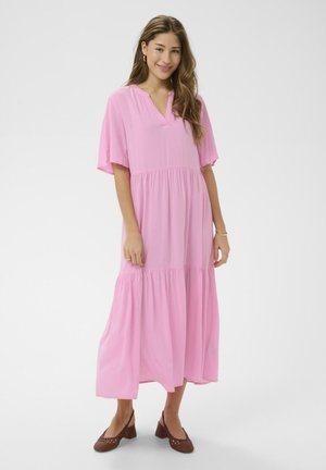 Naeratav naine seisab, kandes lahtist roosat midi-kleit lühikeste varrukatega ja pruune madala kontsaga punutud kingi.