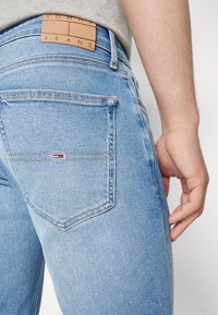 Jeans de mezclilla azul claro con un parche de cuero que dice "TOMMY JEANS", con bolsillos traseros y detalles de costura visibles.