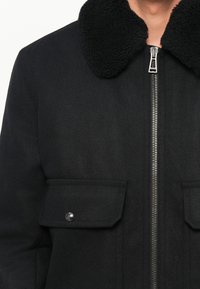 Chaqueta de mezcla de lana negra con cuello de borreguillo, cremallera frontal y dos bolsillos en el pecho con cierres a presión. Textura suave y diseño estructurado.