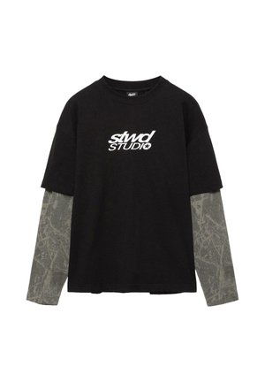 Tricou negru cu mâneci scurte purtat peste o cămașă gri cu mâneci lungi, având un model abstract subtil și inscripția albă „stwd STUDIO” pe piept.