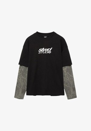 Tricou negru cu mâneci scurte purtat peste o cămașă gri cu mâneci lungi, având un model abstract subtil și inscripția albă „stwd STUDIO” pe piept.