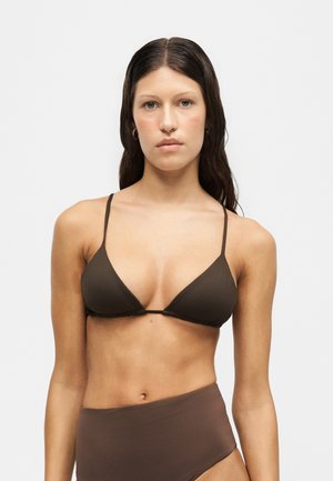 Jeune femme aux longs cheveux foncés portant un haut de bikini triangulaire marron et un bas de bikini taille haute, debout devant un fond blanc uni.