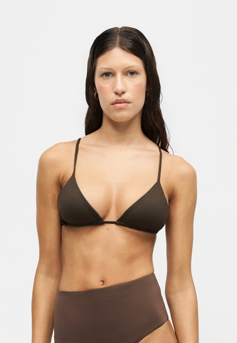 Jeune femme aux longs cheveux foncés portant un haut de bikini triangulaire marron et un bas de bikini taille haute, debout devant un fond blanc uni.