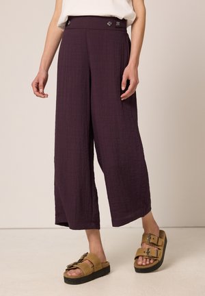 Vrouw draagt losse donkerpaarse wijde broek met knoopsluiting in de taille, wit topje en beige platformsandalen met dubbele gesp.