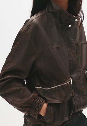 Femme portant une veste en cuir marron foncé avec un col montant, des poches zippées et des poignets élastiqués, les mains dans les poches de la veste.