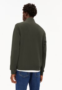 Dunkelgrüner Sweatshirt mit hohem Kragen, gerippten Bündchen und Saum. Hergestellt aus einem glatten Stoff, getragen über einem weißen Hemd mit blauen Jeans.